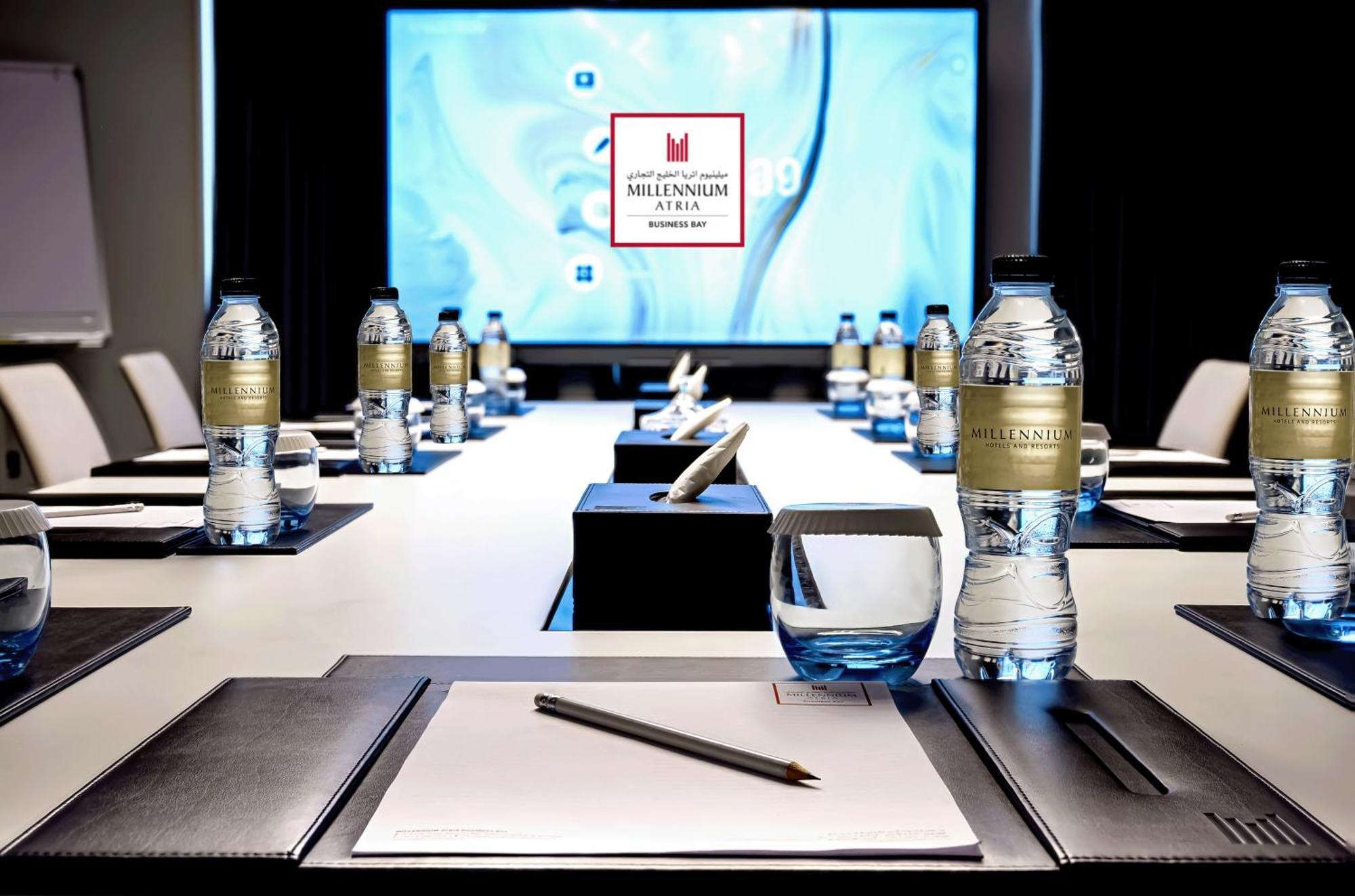 Millennium Atria Business Bay Aparthotel Dubai Exterior photo The photo shows a modern conference room setup. A long table is laid out with neatly arranged stationery, including notebooks and pens, at each seat. Several bottles of water are placed on the table. In the background, a large screen is displayed, li