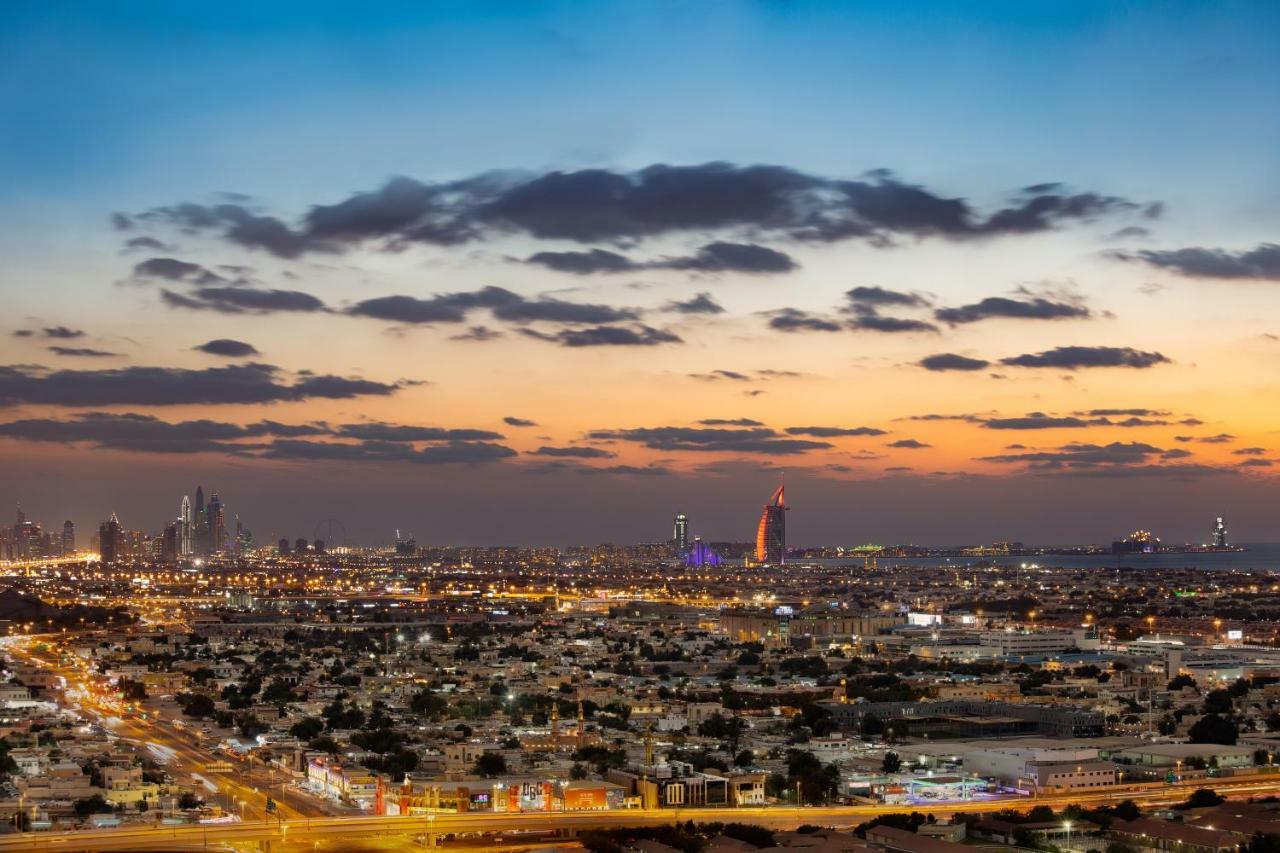 Millennium Atria Business Bay Aparthotel Dubai Exterior photo The photo shows a stunning cityscape at dusk, with a panoramic view of a city skyline. The sky is transitioning from blue to orange, indicating sunset, with scattered clouds creating a colorful backdrop. In the foreground, you can see a sprawling urb