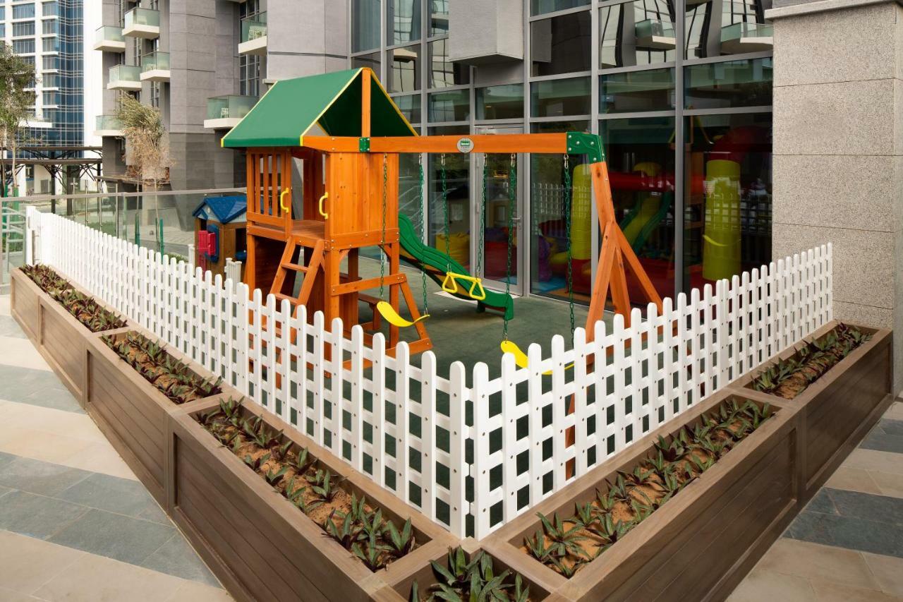 Millennium Atria Business Bay Aparthotel Dubai Exterior photo The image shows a children's playground area, which is enclosed by a white picket fence. The playground features a wooden play structure with a slide, swings, and climbing elements. Surrounding the play area are planters with greenery, and the backdr