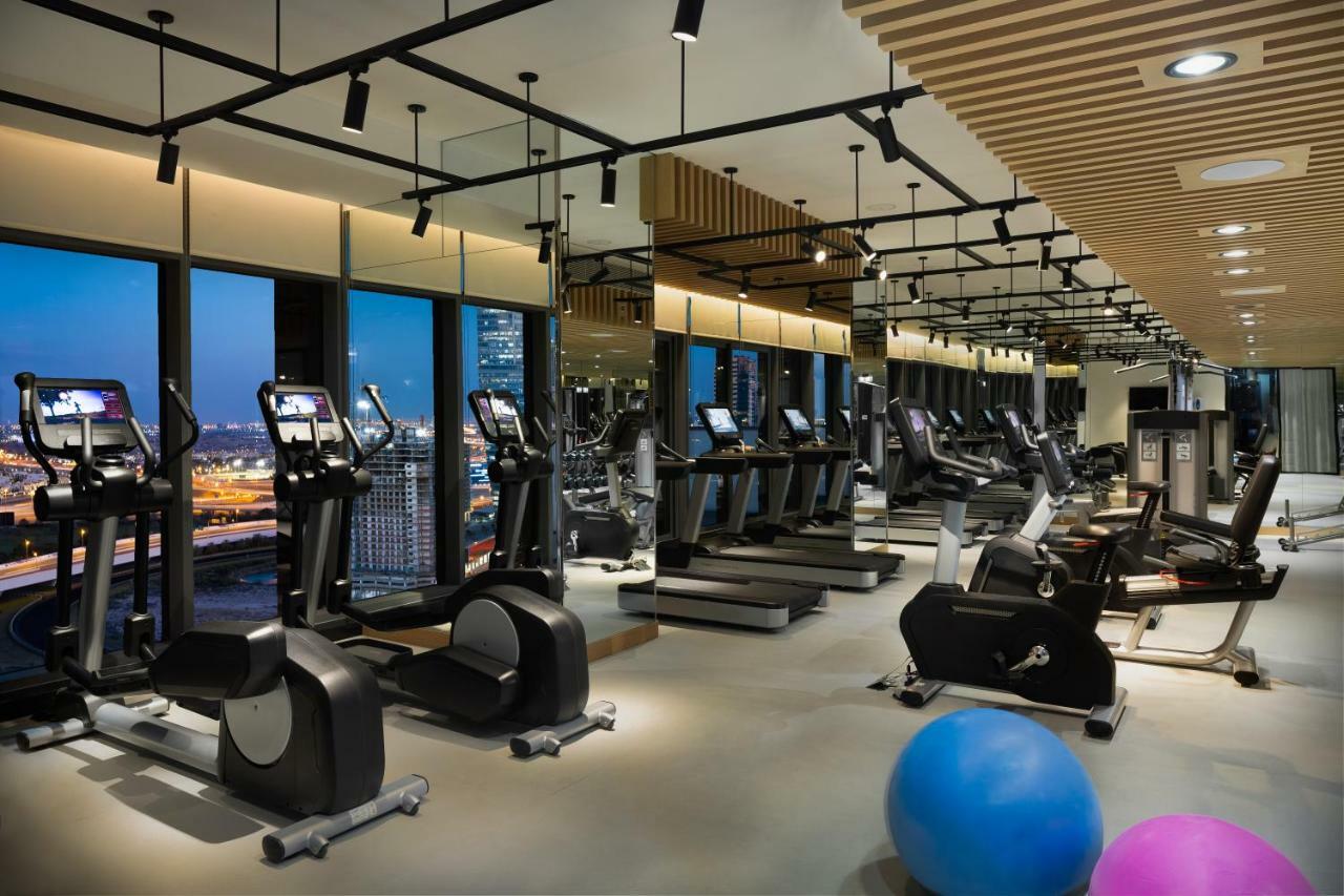 Millennium Atria Business Bay Aparthotel Dubai Exterior photo The photo shows a modern gym with various types of exercise equipment available. There are several treadmills and elliptical machines arranged in rows, with large windows in the background providing a view of the city. The lighting is bright and cont